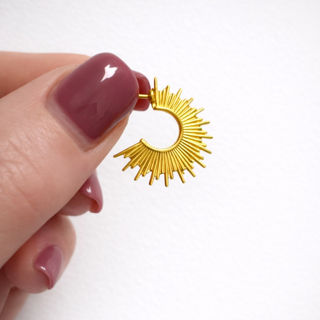 Gold sun-shaped earring held by a hand with maroon nail polish on a white background
