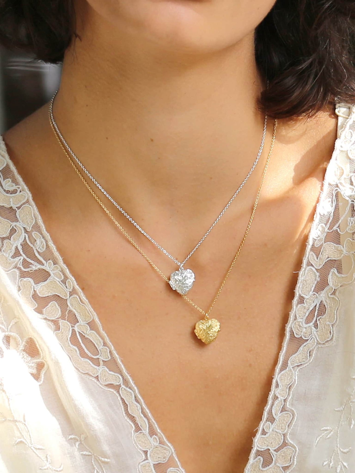 Close-up of a person wearing two necklaces with heart pendants, one gold and one silver, on a blurred background.