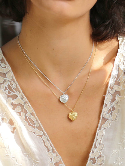 Close-up of a person wearing two necklaces with heart pendants, one gold and one silver, on a blurred background.