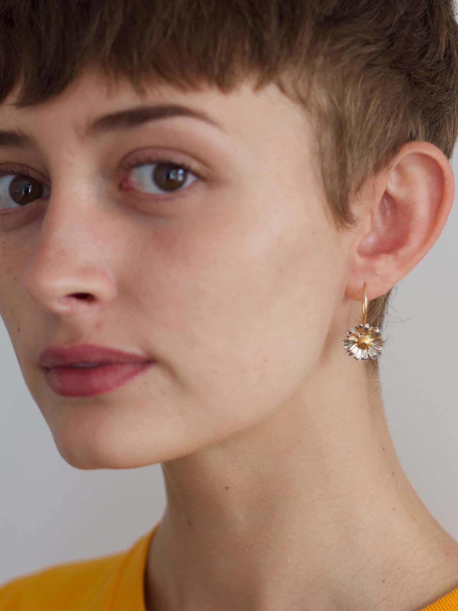 Close-up of a woman wearing an Alex Monroe 22 carat Gold Vermeil & Sterling Silver Big Daisy Hook Earrings with a clear gemstone on a neutral background