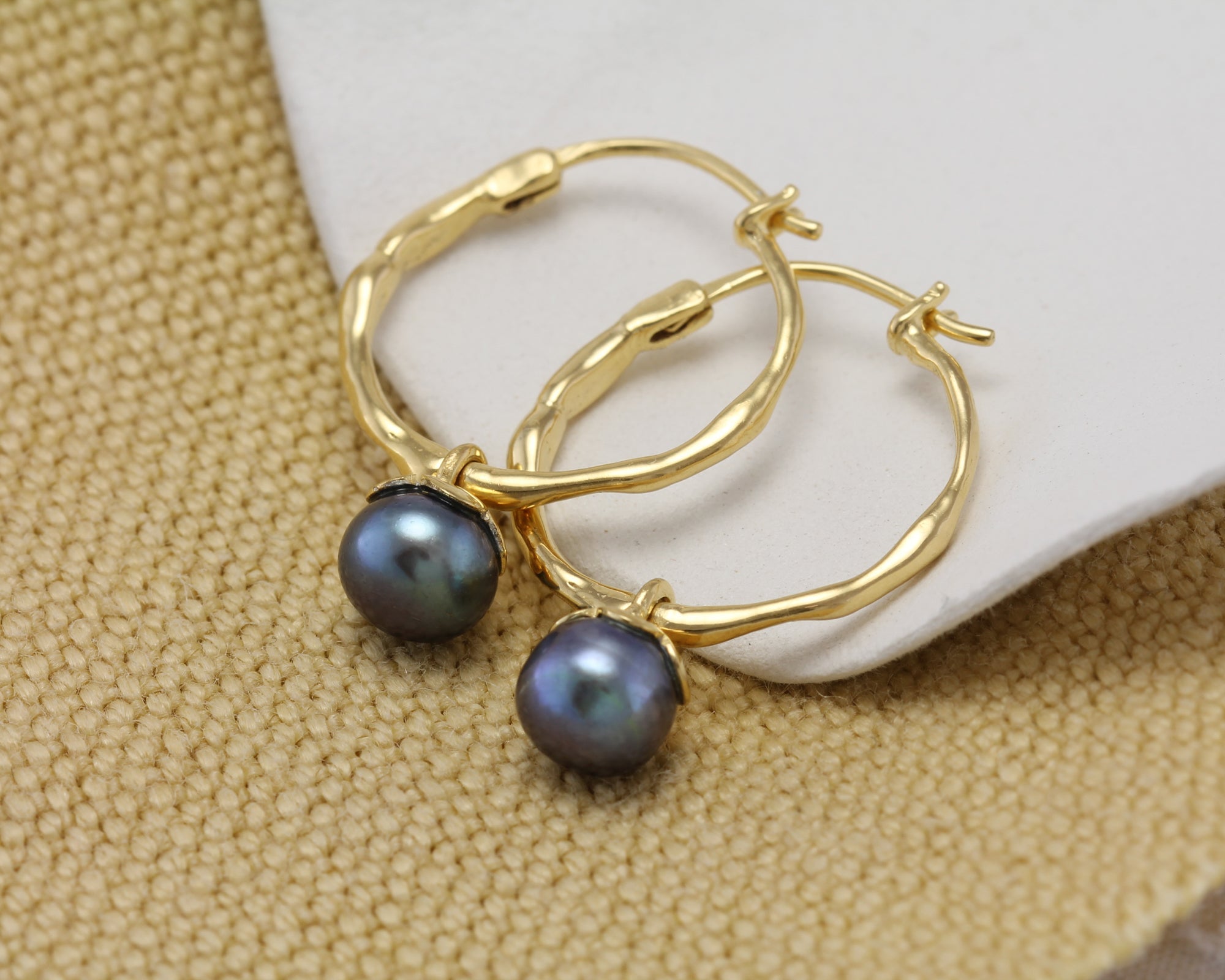 Gold hoop earrings with black pearls on a textured beige surface