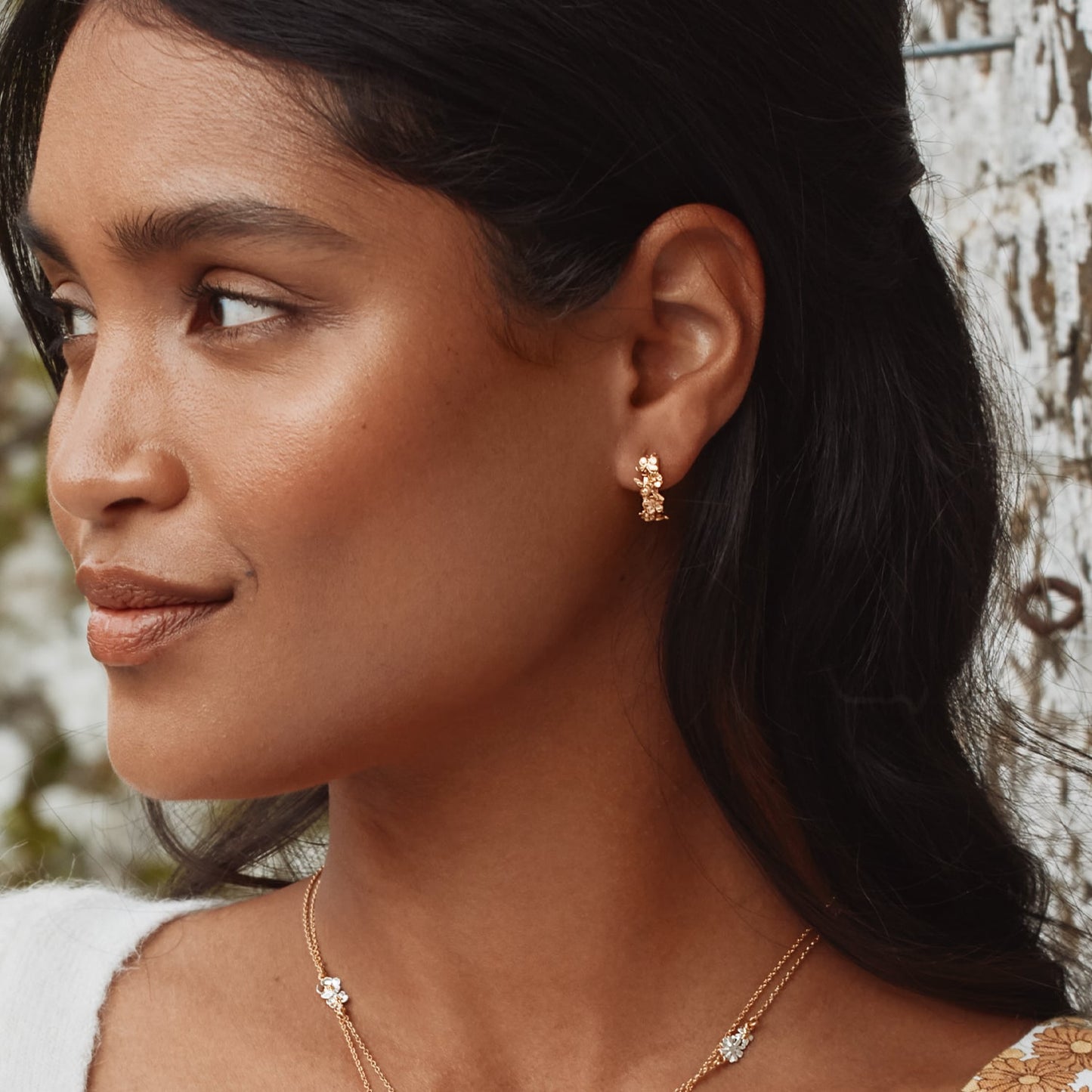 Close-up of a woman wearing gold hoop earrings and a necklace with a blurred natural background