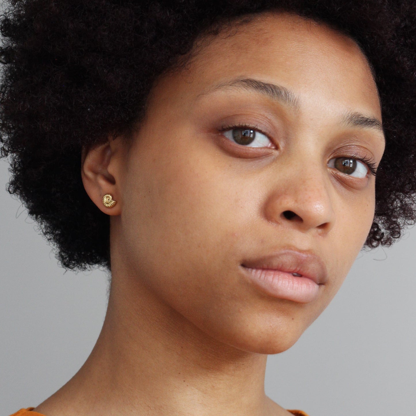 Close-up of a woman with an afro, wearing Alex Monroe Sterling Silver Ammonite Stud Earrings against a gray background