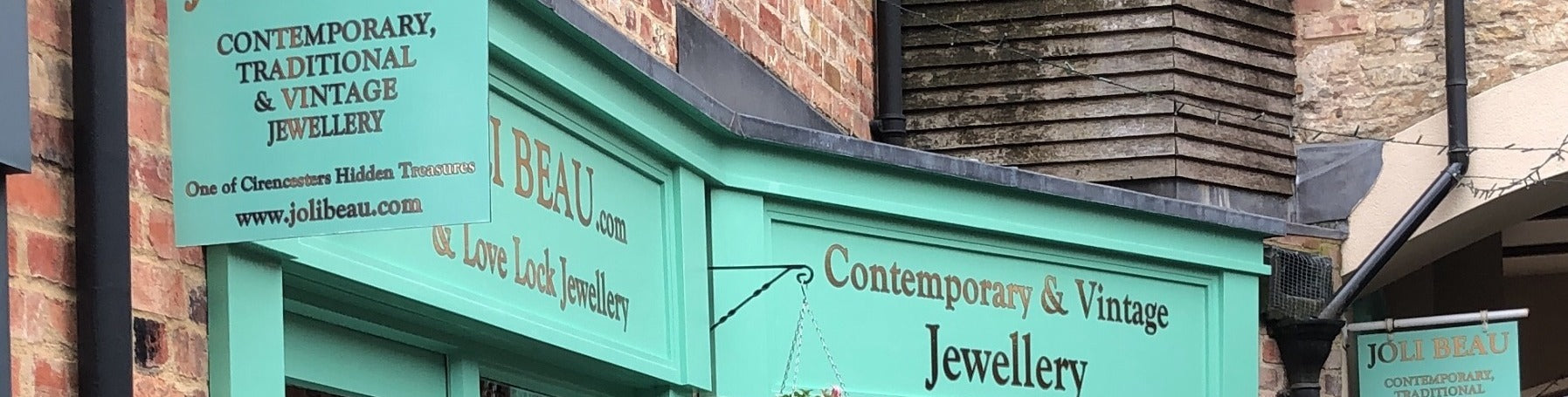 Green storefront sign for Joli Beau jewelry store with text on a brick building.