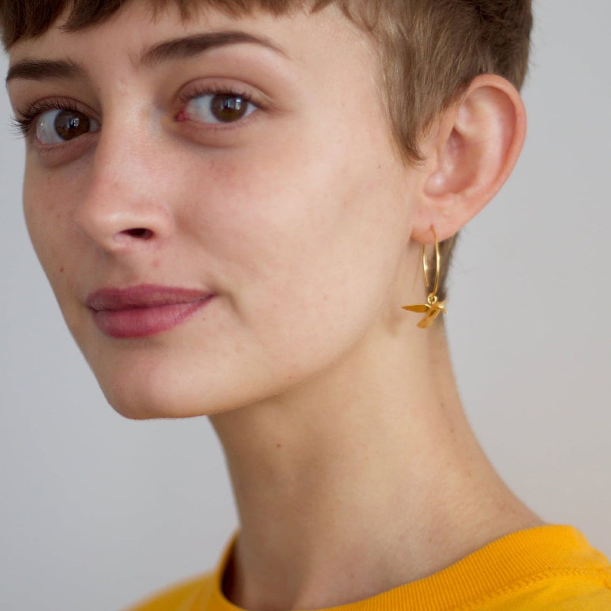 Close-up of a person wearing gold hummingbird hoop earrings with a neutral background 