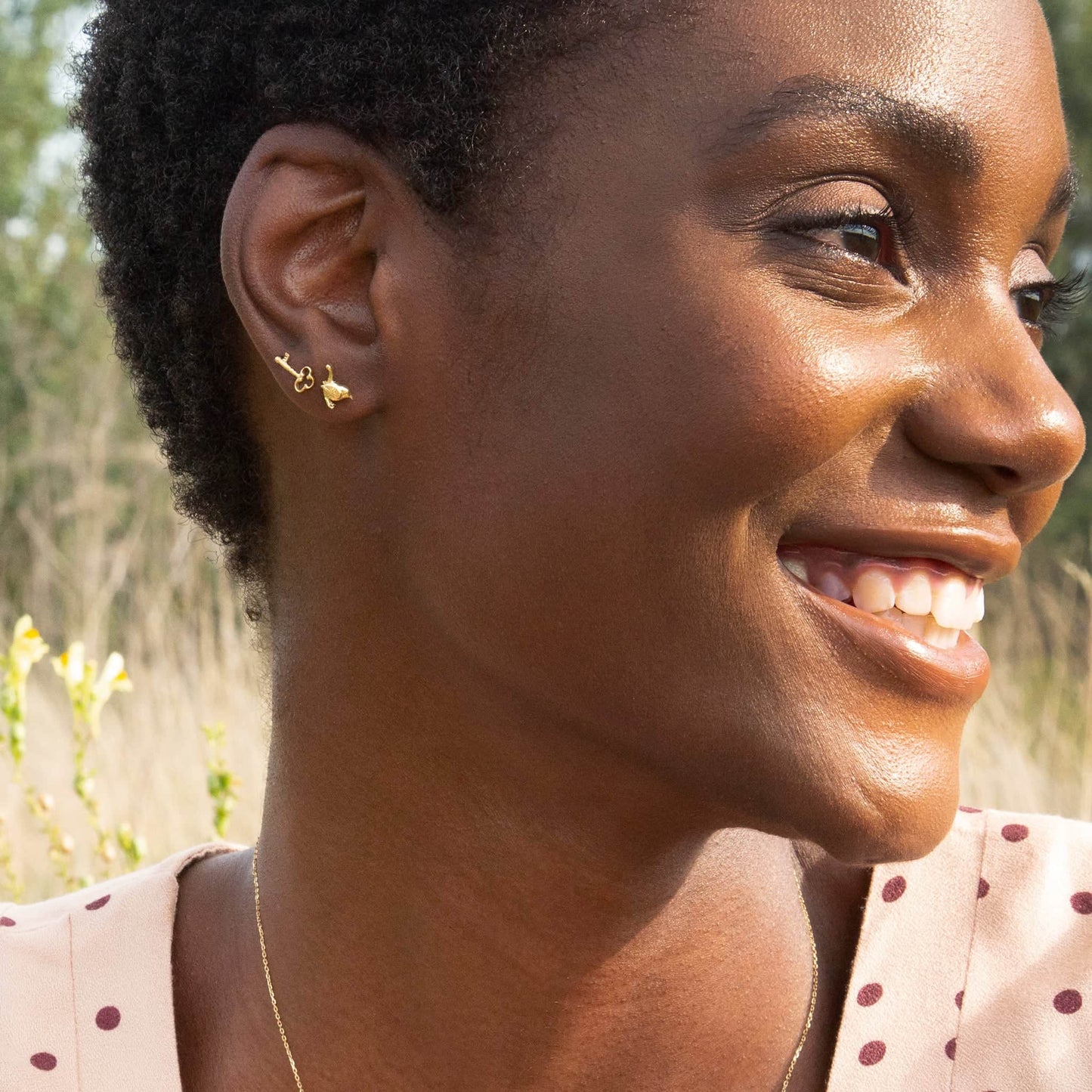 Woman wearing Alex Monroe 18 carat Yellow Solid Gold Teeny Tiny Wren Single Stud Earring with a blurred natural background