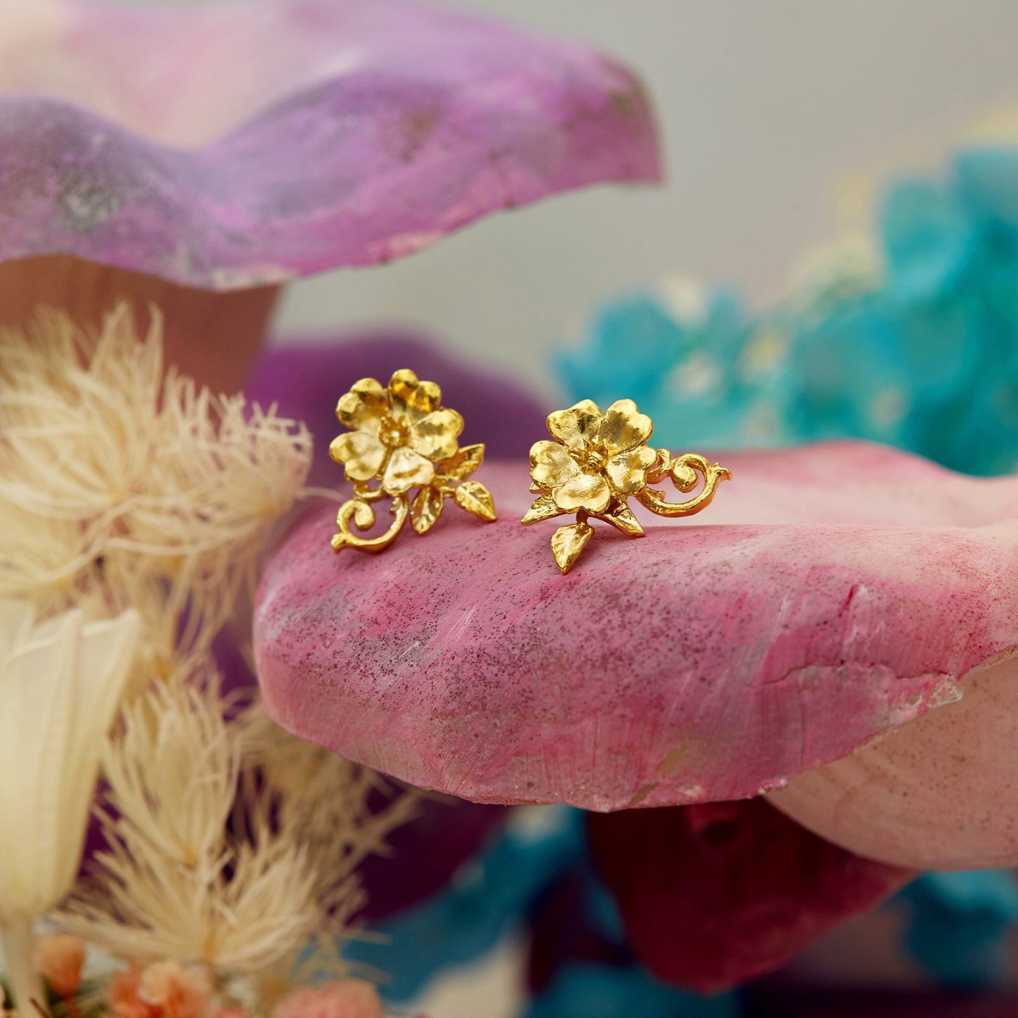 Alex Monroe 22 carat Gold Vermeil Ornate Rose Stud Earrings on a pink stone with colorful flowers in the background